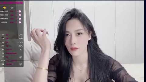 Media: Video of a young East Asian woman with long black hair, fair skin, and light makeup, wearing a sheer black top, sitting on a white couch, with a smartphone screen showing social media stats in the background.