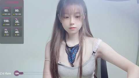 Media: A video of an East Asian woman with long, straight brown hair, wearing a beige ribbed tank top, and a black and blue floral necklace. She has a pale complexion, small breasts, and a neutral expression. Background shows a simple, light-colored room.