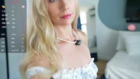 Media: Video of a blonde woman with fair skin and light makeup, wearing a white off-shoulder top and pearl necklace. Background shows a modern living room with a TV and white sofa.