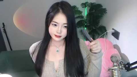 Media: Video of a young East Asian woman with long black hair, wearing a beige cardigan and necklace, speaking into a microphone in a cozy room with a green couch and potted plant.