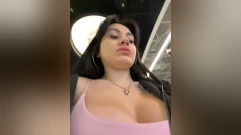 Media: A video of a Latina woman with long black hair, wearing a lavender spaghetti-strap top that reveals ample cleavage, large hoop earrings, and a silver necklace. She stands in a modern, well-lit room with a round ceiling light.