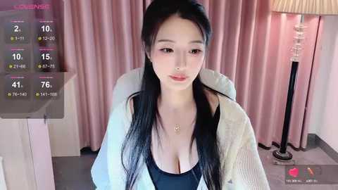 Media: Video of a young Asian woman with long black hair, wearing a black top, white cardigan, and a necklace, seated in a room with pink curtains and a lamp.