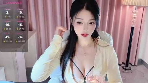 Media: Video of an East Asian woman with long black hair, wearing a cream hoodie and black bra, sitting in a room with pink curtains and a lamp.