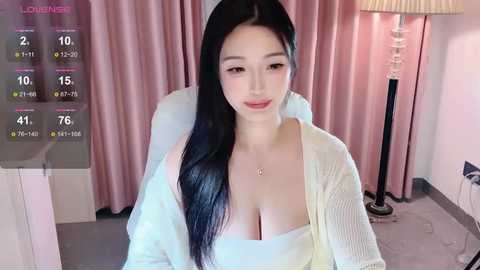 Media: A video of an Asian woman with long black hair and fair skin, wearing a white top that reveals cleavage, sitting in a room with pink curtains and a lamp.