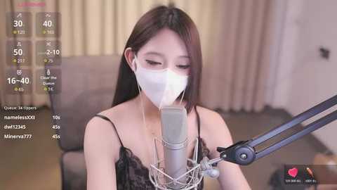 Media: A video of an Asian woman with long black hair, wearing a white face mask and a black lace top, singing into a microphone. Background features beige curtains and a grey chair.