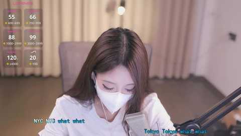 Media: Video of an Asian woman with long brown hair, wearing a white face mask, working at a desk with a microphone. Background shows beige curtains and a lamp.