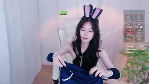 Media: Video of a slender East Asian woman with long black hair, wearing a black top and blue bunny ears, adjusting a blue dress. Background features a white room with a plant.