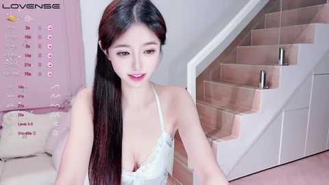 Media: A video of an East Asian woman with long black hair and fair skin, wearing a white lace bra, sitting on a white couch in a modern, minimalist room with a calendar and staircase in the background.