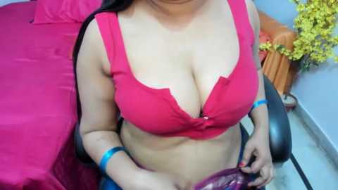 Media: Video of a woman with light brown skin, wearing a red bra that accentuates her large breasts, sitting on a bed with a pink sheet, holding a purple cloth.