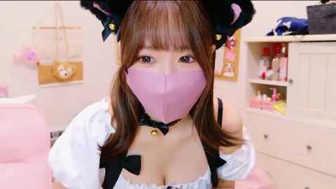 Media: Video of an Asian woman wearing a pink face mask, black hat, and revealing white dress with black ribbon accents, in a pastel-colored room with shelves and plush toys.