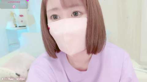 Media: Video of a young Asian woman with bob-cut brown hair and light skin, wearing a white face mask and lavender top, indoors, with a soft-focus background featuring a plush toy and a white wall.