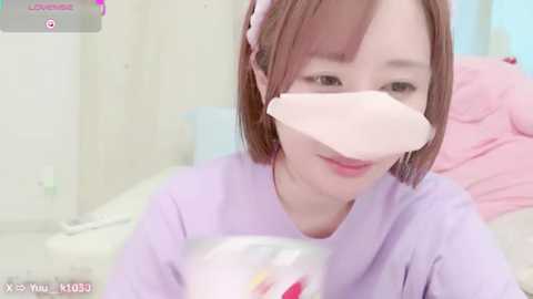 Media: A video of a young Asian woman with short brown hair and light skin, wearing a lavender t-shirt, with a pink surgical mask covering her mouth, smiling. The background is a soft pastel room.