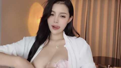 Media: Video of a young East Asian woman with long black hair, fair skin, and light makeup. She wears a white unbuttoned shirt, exposing a pink bra, and a delicate necklace. Background shows a warm, softly lit room with beige curtains.