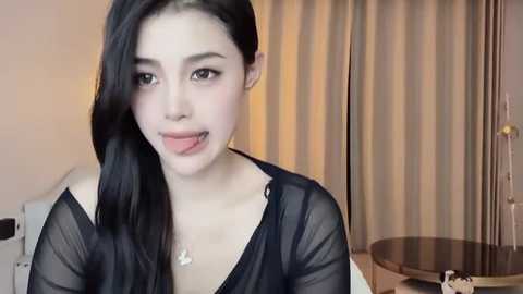 Media: Video of a young Asian woman with long black hair, fair skin, and a playful expression, wearing a sheer black top, in a modern, softly lit room with beige curtains and a round table.