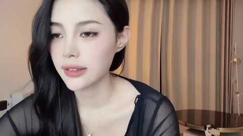 Media: A close-up video of an Asian woman with fair skin, straight black hair, and subtle makeup, wearing a sheer black top, standing in a room with beige curtains and a wooden table.