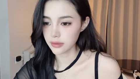 Media: Video of a young East Asian woman with long black hair, pale skin, and large brown eyes, wearing a black choker and sheer black top. She has a neutral expression. Background shows beige curtains and a power outlet.