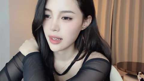 Media: Video of an East Asian woman with fair skin, long black hair, and a slender physique, wearing a sheer black top, sitting indoors near a wooden chair and beige curtains.