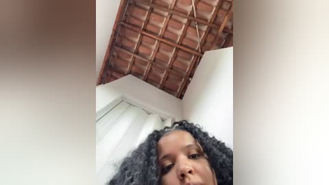 Media: Video of a young woman with dark curly hair, looking up at a rustic, red-tiled roof with a white wall and window in the background.