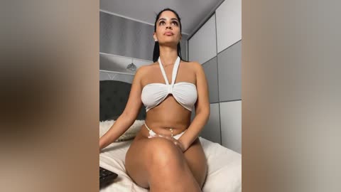 Media: Video of a Latina woman with long black hair, medium skin tone, sitting on a bed in a modern bedroom, wearing a white halter bikini, revealing her toned physique and medium-sized breasts.