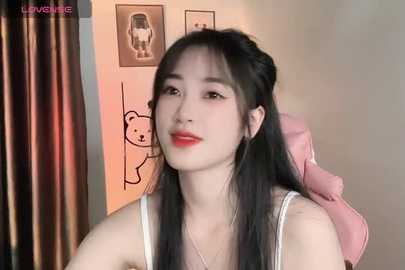 Media: Video of an East Asian woman with long black hair, wearing a white tank top, sitting in a pink gaming chair. Background features a black bear illustration on beige walls.