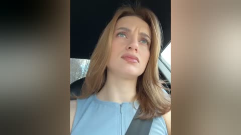 Media: Video of a young woman with light skin and shoulder-length brown hair, wearing a light blue top, seated in a car with a silver seatbelt. She has a contemplative expression, gazing out the window.