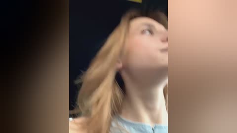 Media: Video of a woman with fair skin and long blonde hair, wearing a light blue top, seen from the side. The background is dark and blurry, making the subject stand out.