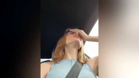 Media: Video of a young woman with shoulder-length blonde hair, wearing a light blue sleeveless dress, sitting in a car. She is covering her eyes with her right hand, appearing distressed. The car's interior is dimly lit, with a black ceiling and a bright window on the right.