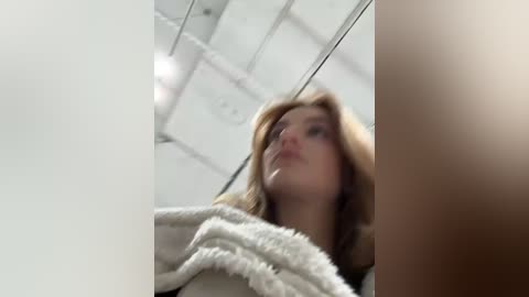 Media: Video of a young woman with fair skin and blonde hair, wearing a beige, fluffy robe, standing in a bright, modern room with white ceiling tiles and recessed lights.