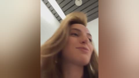 Media: A blurry, out-of-focus video of a blonde woman with fair skin and light makeup, seen from below, standing indoors with a modern, gray ceiling.