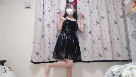 Media: A video of an East Asian woman in a black, shiny latex dress, wearing red cat ears, a white face mask, and standing against a patterned curtain in a dimly lit room.