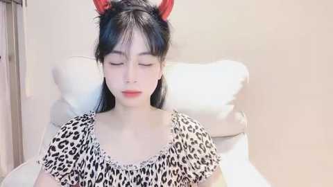 Media: Video of an East Asian woman with long black hair, wearing a leopard print top and red cat ears, lying on a white pillow in a soft, neutral-toned room.