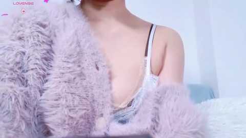 Media: Video of a woman with light skin, wearing a sheer, white lace bra and a fluffy, lavender robe, partially covering her breasts. Background features a white bed with a white, textured blanket.