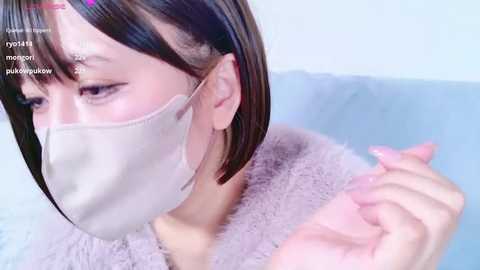 Media: Video of an East Asian woman with straight, shoulder-length brown hair, wearing a beige face mask and a fluffy, light pink robe. She is gently touching her face with her left hand. Background is a soft blue.