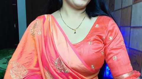 Media: Video of a woman with fair skin, medium build, wearing a vibrant orange sari with intricate gold embroidery, adorned with a black necklace, against a dark background.