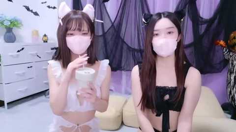 Media: Two young Asian women in Halloween-themed lingerie, white cat ears, and face masks, sitting on a sofa in a room with purple curtains and bat decorations.