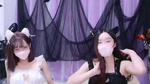 Media: Video of two Asian women in Halloween costumes: one with white cat ears and a white dress, the other with black cat ears and a black dress, both wearing white face masks. Background features purple and black drapes.