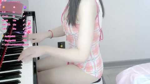 Media: Video of an Asian woman with long black hair, wearing a pink plaid dress, playing a piano in a modern, minimalistic room with white walls and a bed in the background.