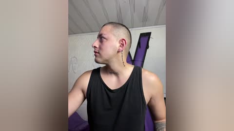 Media: Video of a young man with a shaved head, wearing a black sleeveless tank top and long earrings, standing against a white wall with a purple couch behind him.