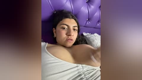 Media: A video of a Latina woman with medium brown skin and dark hair lying on a purple tufted leather headboard, wearing a white off-shoulder top, with a blurred background.