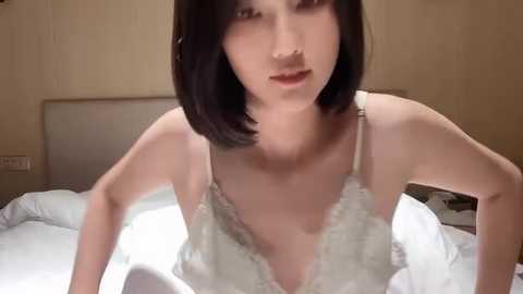 Media: Video of a young Asian woman with straight, shoulder-length black hair, wearing a white lace bra, leaning forward on a bed in a dimly lit, beige-walled bedroom.