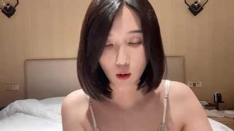 Media: Video of a young Asian woman with straight, shoulder-length black hair, fair skin, wearing a white spaghetti-strap top, eyes closed, and mouth slightly open in a bedroom with beige walls and a bed.