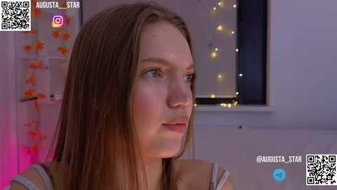 Media: Video of a young Caucasian woman with straight, light brown hair, wearing a brown top, in a dimly lit room with fairy lights and floral decor.