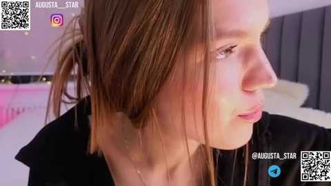 Media: Video of a fair-skinned young woman with straight, light brown hair, wearing a black top, captured in a dimly lit bedroom with a gray upholstered headboard.