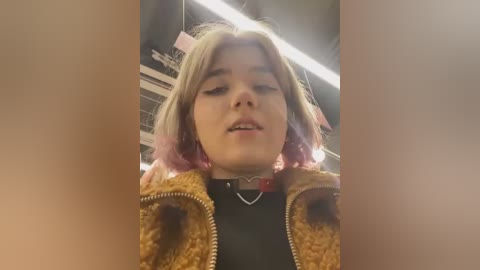 Media: A video of a young woman with light skin and pink-streaked hair, wearing a black shirt and a brown, furry jacket with a heart-shaped necklace, taken from a low angle in a dimly lit room.