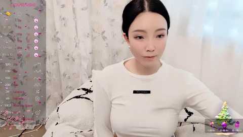 Media: Video of a young East Asian woman with fair skin and straight black hair, wearing a white long-sleeved shirt, seated indoors. Background features white floral-patterned curtains and a digital screen displaying a chat interface.