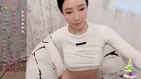 Media: A video of an East Asian woman with a slim build, fair skin, and short black hair, wearing a white crop top, sitting in a cozy room with a floral-patterned wall and a virtual Christmas tree in the corner.