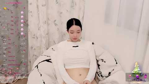 Media: Video of an East Asian woman with pale skin, dark hair in a bun, wearing a white crop top and oversized jacket, sitting on a white beanbag chair in a minimalistic room with white curtains and a calendar on the left.