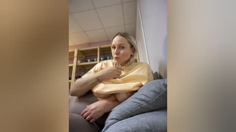 Media: A video of a blonde woman with fair skin, wearing a yellow t-shirt lifted to reveal her bare breasts, sitting on a gray couch in a room with a white ceiling and bookshelves.