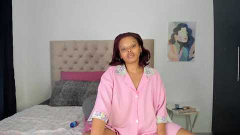 Media: Video of a Black woman with medium skin tone, wearing a pink robe, sitting on a bed with gray bedding, white walls, and a colorful abstract painting.