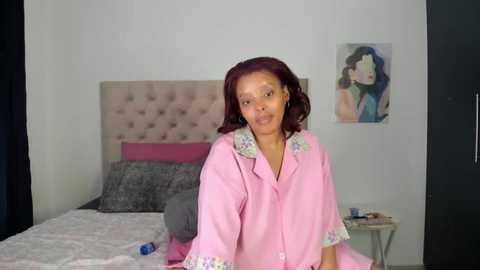 Media: Video of a smiling Black woman with medium skin tone and curly dark hair, wearing a light pink robe with floral accents, standing in a minimalist bedroom with a beige tufted headboard and abstract art on the wall.